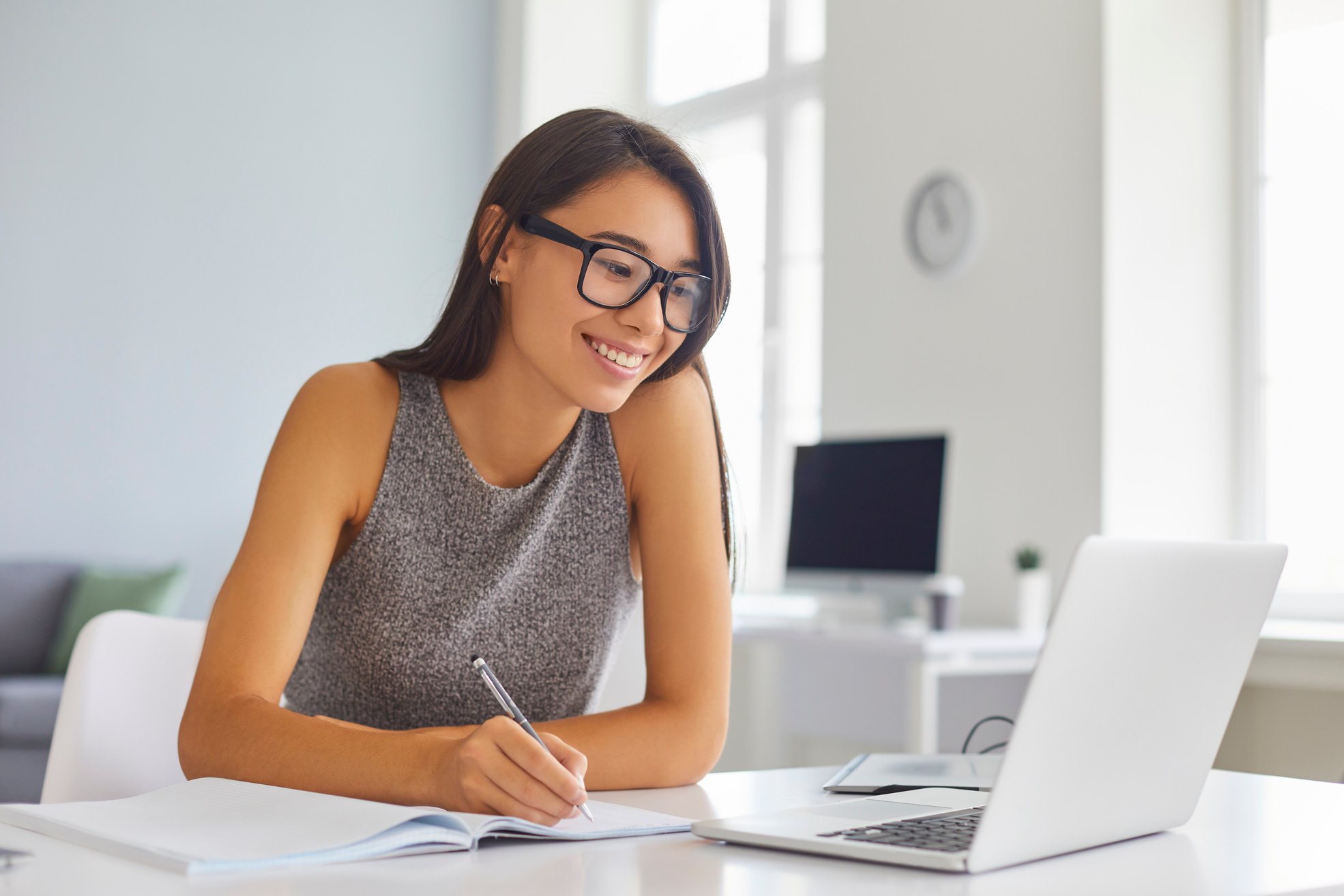 Smiling Woman Office Worker Sitting at Desk, Looking at Laptop Screen and Making Notes during Online Conference